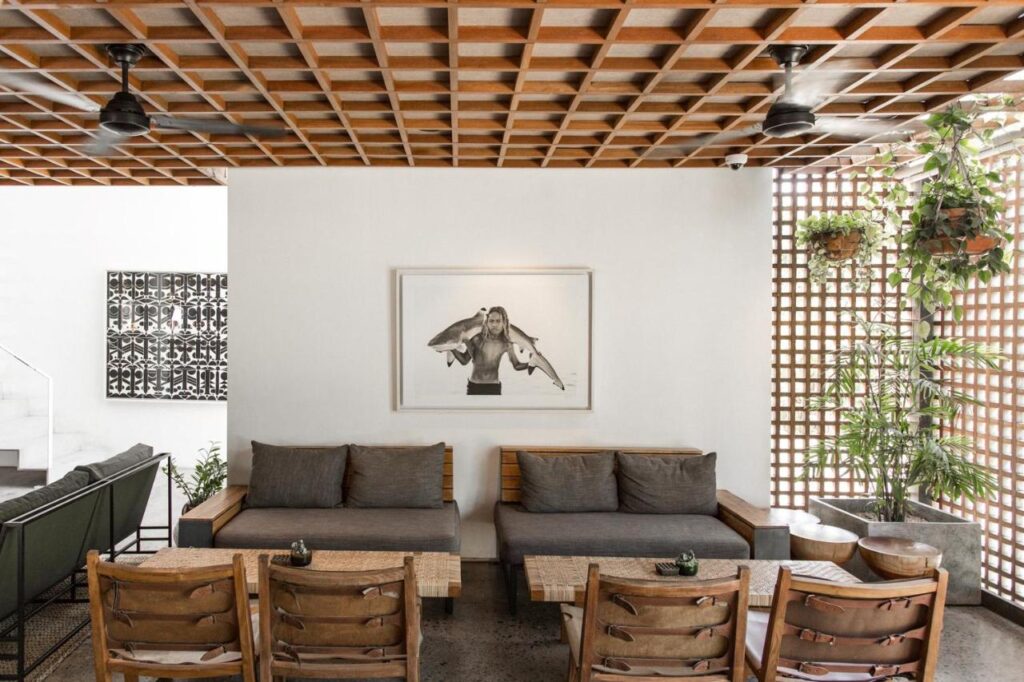14 UNIQUE Boutique Hotels in Canggu, Bali 25 Modern lounge with wooden furniture, gray cushions, potted plants, and a wooden grid ceiling. Reminiscent of boutique hotels in Canggu, a black-and-white artwork hangs above sofas, creating a stylish, minimalistic atmosphere.