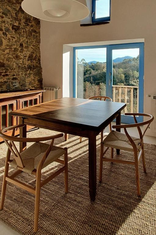 A wooden dining table with three chairs sits on a woven rug near a window with a scenic view of trees and mountains, reminiscent of boutique hotels in Asturias. Sunlight streams through the window, casting shadows on the table and floor.