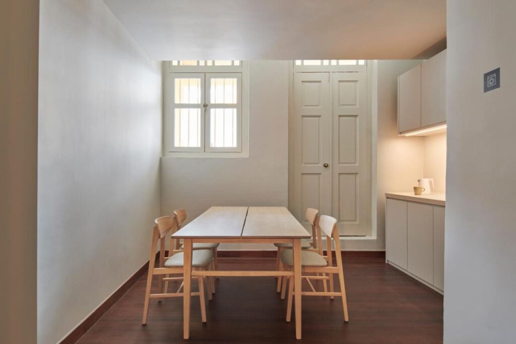 13 Most Unique Hotels in Singapore for an Incredible Holiday 9 Minimalist dining area with a wooden table and four chairs on a dark wood floor, set against light-colored walls—reminiscent of the stylish interiors often found in unique hotels in Singapore. Tall windows and a kitchenette complete the inviting space.