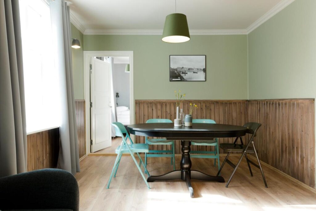 Where to Stay in Reykjavík: Top Guide 20 A dining room with light green walls, wooden wainscoting, and a black oval table is perfect inspiration for those searching where to stay in Reykjavik—three turquoise chairs, one black chair, pendant lamp, and sunlight streaming through the window.