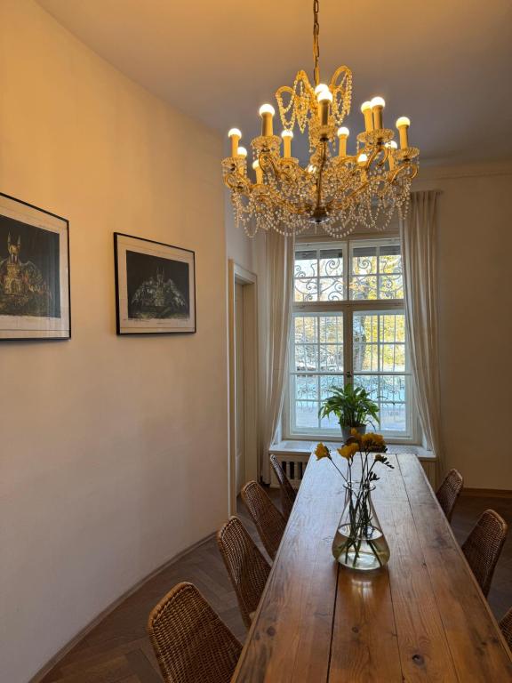 FAIRYTALE Castle Hotels in Bavaria 6 A cozy dining room reminiscent of castle hotels in Bavaria, with a wooden table, wicker chairs, a glass vase of yellow flowers, framed art on the wall, sheer curtains at the window, and an ornate chandelier illuminating the space.