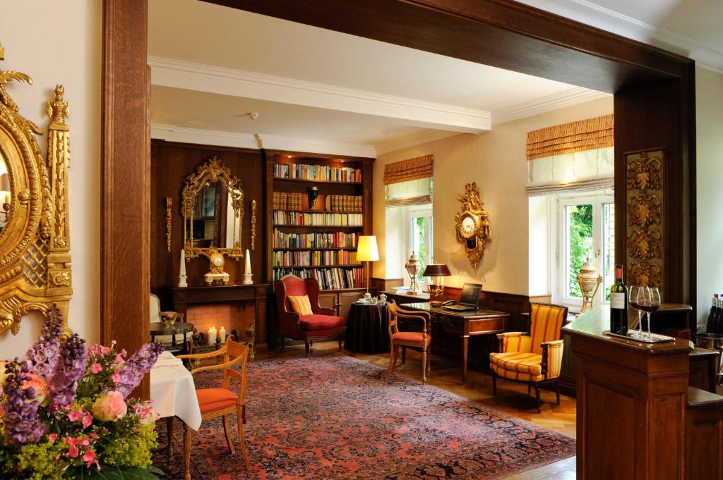 Authentic Boutique Hotels in Munich 16 Elegant living room with antique furniture, bookshelves, a fireplace, ornate mirrors, a patterned rug, and fresh flowers—reminiscent of the charm found in boutique hotels in Munich. Large windows fill the cozy reading nook and desk with natural light.