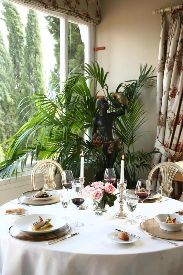 25 Weekend Trips from Madrid Max. 2h Away 8 A round dining table set for four with elegant plates, glassware, wine glasses, and candles evokes the charm of weekend trips from Madrid. Pink roses grace the center, framed by lush plants and a classical statue by a large window.