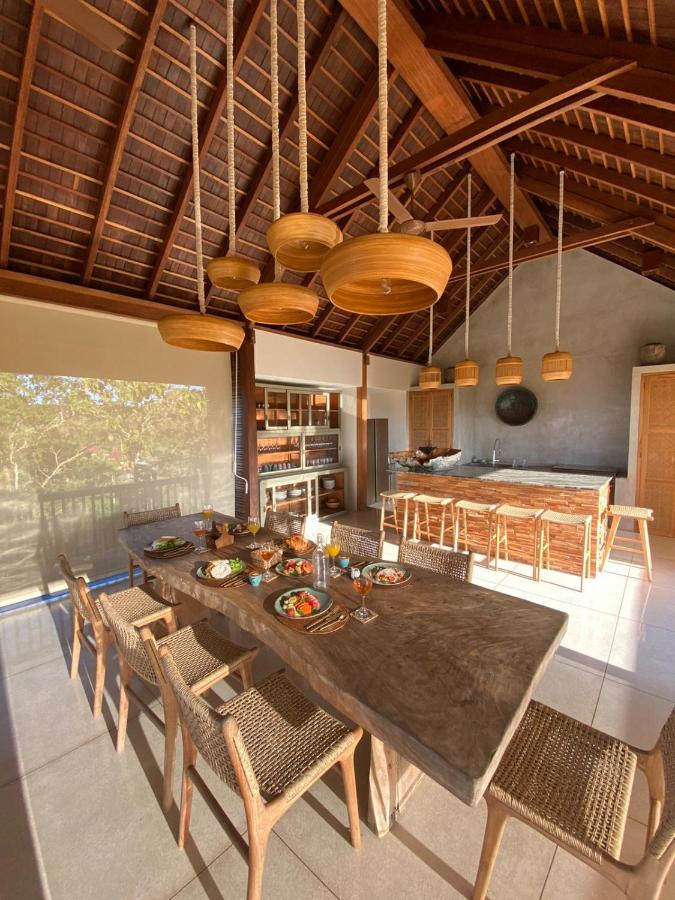 A sunlit, modern kitchen and dining area—hallmarks of the best family villas in Bali—with a wooden dining table set for a meal, woven chairs, pendant lights, high wooden ceiling, and large windows overlooking trees. Kitchen island and cabinets are visible in the background.
