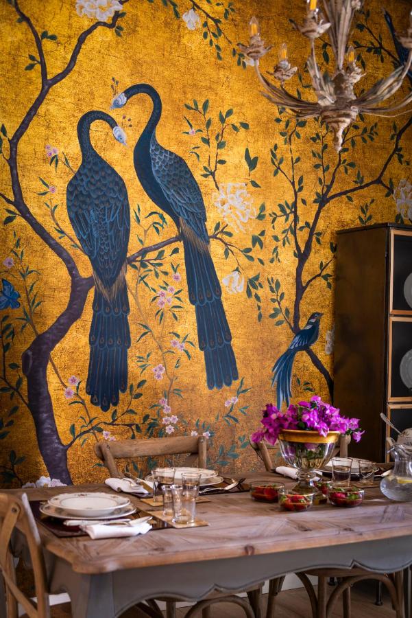 10 UNIQUE Hotels with Pool in Seville 31 A dining room with a wooden table set for a meal, in front of a gold wall mural depicting two blue peacocks on a branch with flowers and a small bird. Reminiscent of the elegance found in hotels with pool in Seville, bright pink flowers decorate the table.