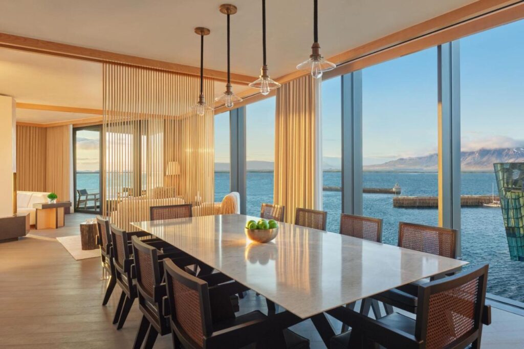 Where to Stay in Reykjavík: Top Guide 5 Modern dining room with a long table and dark chairs, large windows offering sea and mountain views—perfect inspiration for those seeking where to stay in Reykjavik—with pendant lights, warm wood accents, and beige decor throughout.