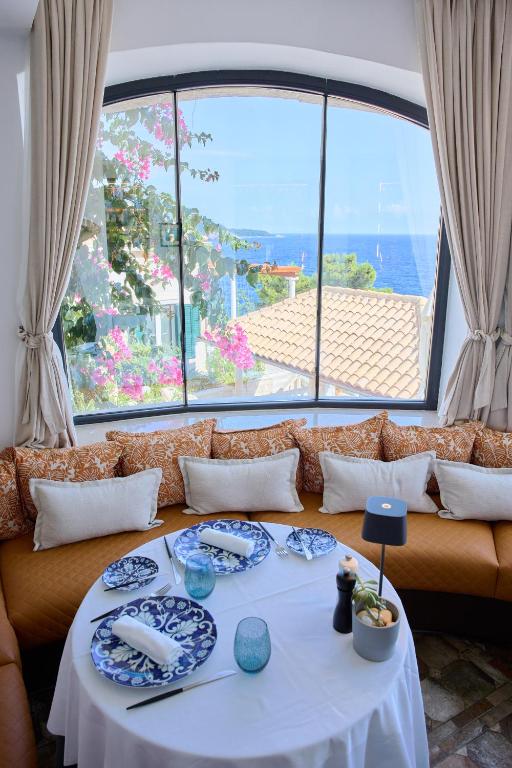 Most Unique Boutique Hotels in Dubrovnik 7 A round table set for four with blue and white plates sits by a curved cushioned bench under a large window, offering a scenic view of the sea, tiled rooftops, and blooming pink flowers—typical of boutique hotels in Dubrovnik.