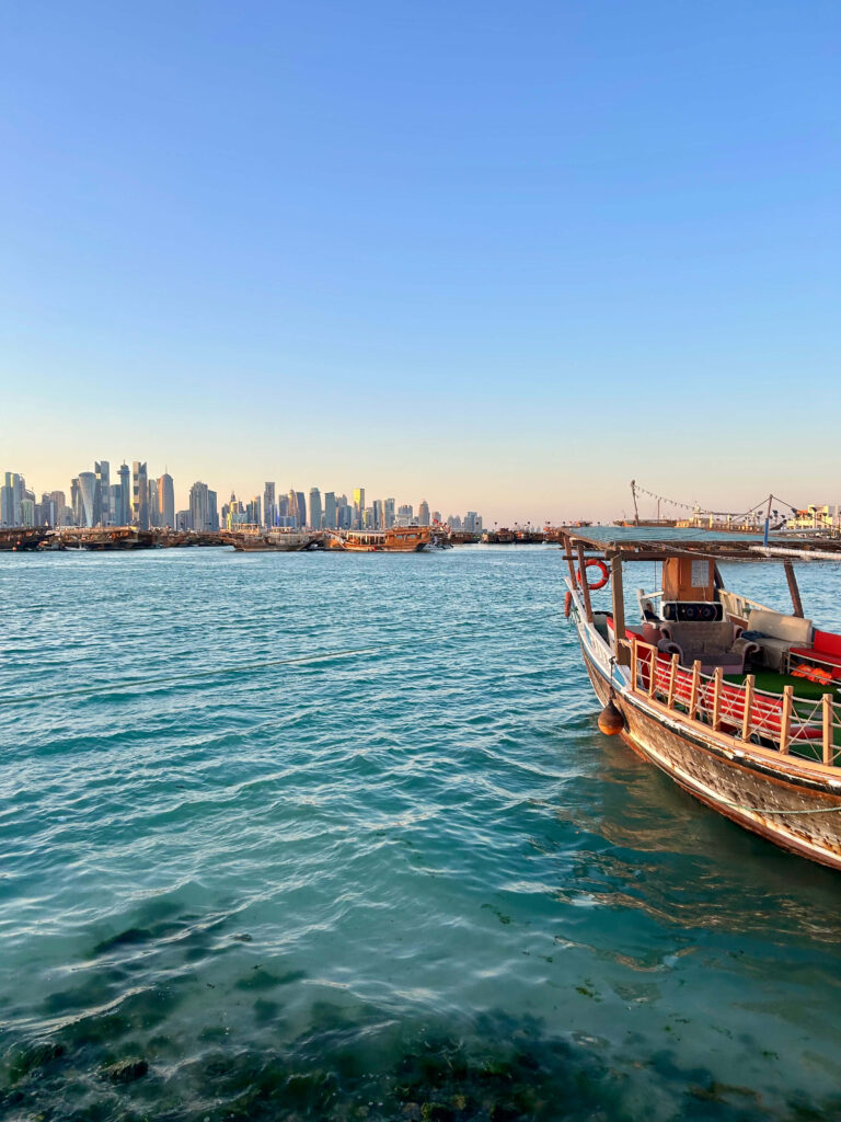 A traditional wooden boat floats on turquoise water with a modern city skyline in the background, capturing the charm of Doha’s contrasts—an inspiring scene for those exploring where to stay in Doha under a clear blue sky.