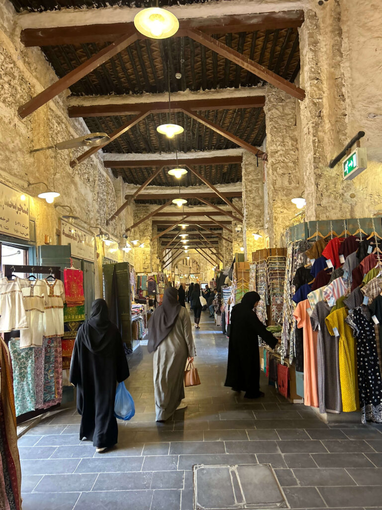 Covered market corridor with stone walls and wooden beams, clothing and fabrics displayed on stalls. Several women in abayas shop under hanging lights. Arabic signs above the shops hint at local life and where to stay in Doha.