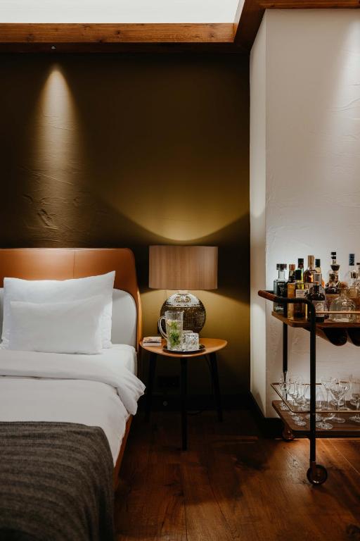 Authentic Boutique Hotels in Munich 20 A cozy bedroom inspired by boutique hotels in Munich features a neatly made bed with white bedding, a bedside table holding a lamp and decorative jar, and a bar cart stocked with bottles and glassware, all set against warm lighting.