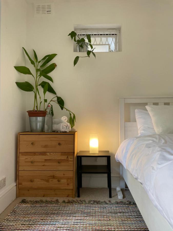 15 Best Boutique Hotels in London, UK 12 A cozy bedroom corner reminiscent of the best boutique hotels London offers, featuring a wooden dresser, a potted plant, a black nightstand with a glowing lamp, and white bedding. A small window with greenery completes the inviting space.