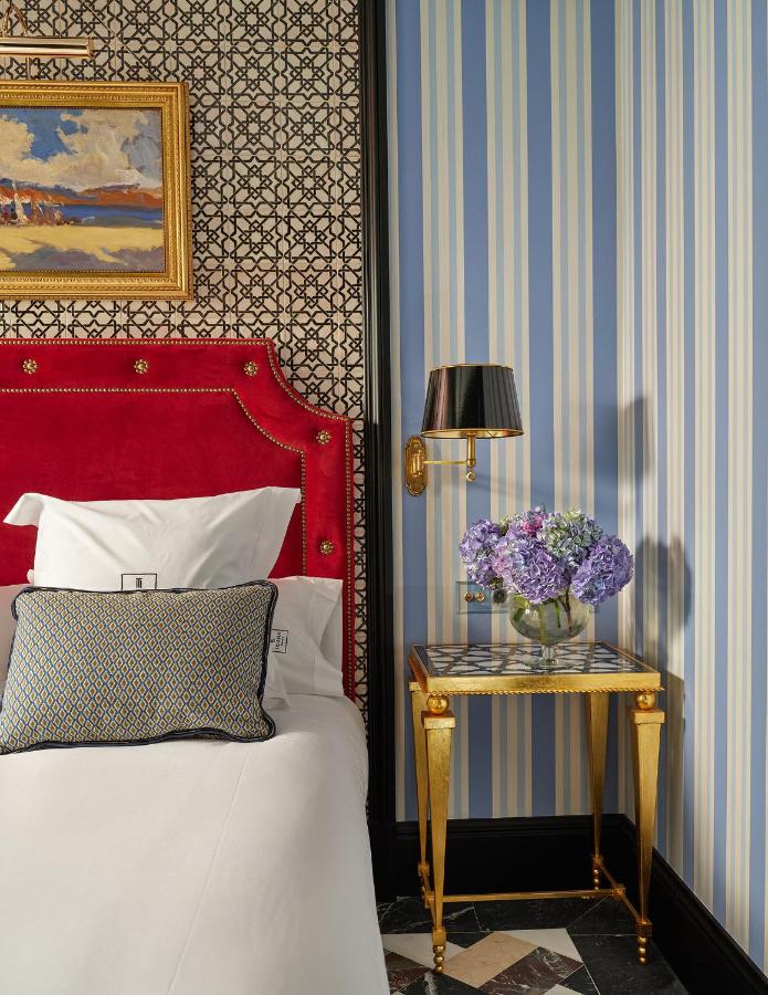 1 day in Seville Spain: My Favorite City 11 A stylish bedroom with a red velvet headboard, patterned wallpaper, and a side table holding a vase of purple flowers evokes the elegance you'd experience during 1 day in Seville, Spain. Blue and white striped walls and art complete the look.