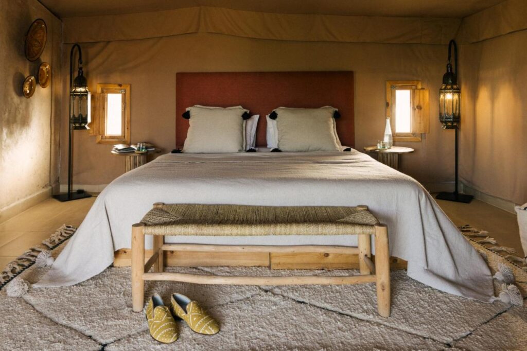 8 SPECTACULAR Agafay Desert Camps 19 A cozy bedroom inspired by Agafay Desert camps, with a large bed, two pillows, a woven bench at the foot, yellow slippers on a rug, and soft lantern lighting. Small windows and decorative items adorn the walls.