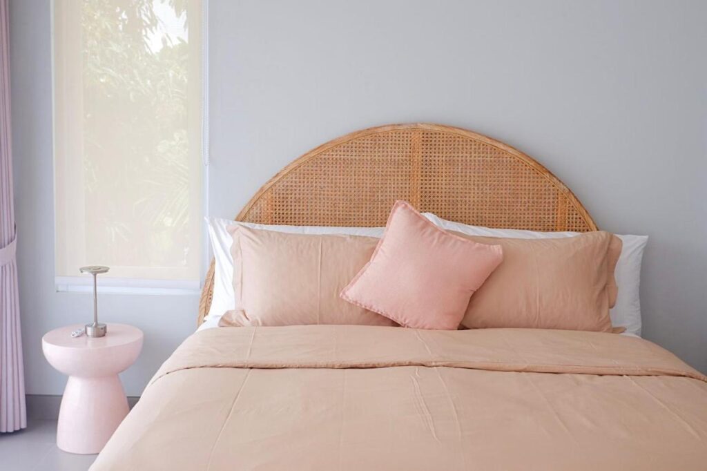 14 UNIQUE Boutique Hotels in Canggu, Bali 30 A neatly made bed with light peach bedding and a rattan headboard, featuring a matching pillow, sits beside a round pink nightstand with a lamp—evoking the serene style of boutique hotels in Canggu—in a softly lit, minimalist bedroom.
