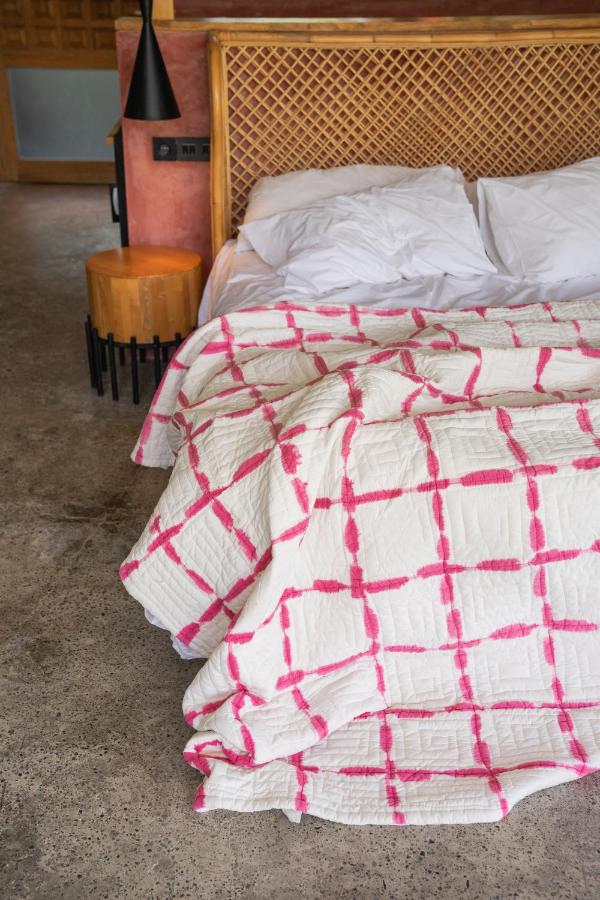 14 UNIQUE Boutique Hotels in Canggu, Bali 18 A bed with white pillows and a quilt featuring a pink grid pattern, slightly rumpled at the foot—just like those found in boutique hotels in Canggu. A wicker headboard and wooden nightstand with a black lamp sit on a concrete floor.
