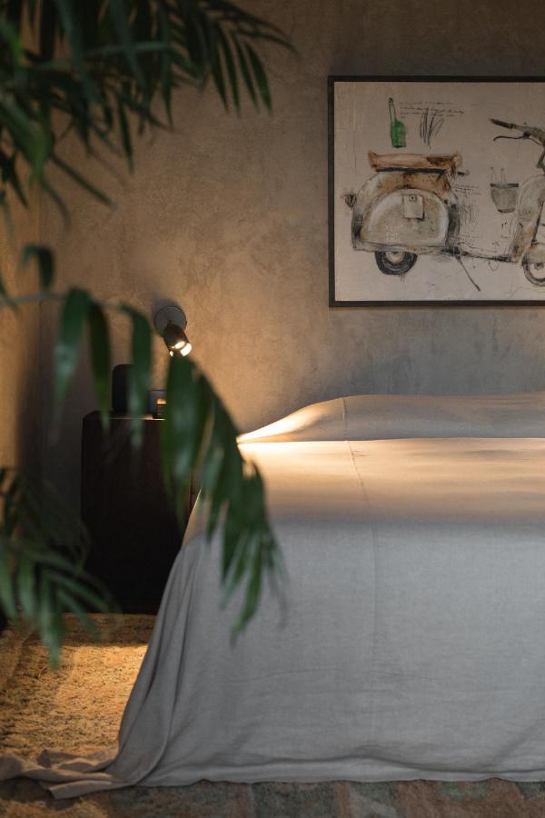 14 UNIQUE Boutique Hotels in Canggu, Bali 3 A cozy bedroom reminiscent of boutique hotels in Canggu, featuring a neatly made bed with a light gray blanket, a small bedside table with a lamp, a leafy plant in the foreground, and vintage scooter wall art above the bed.