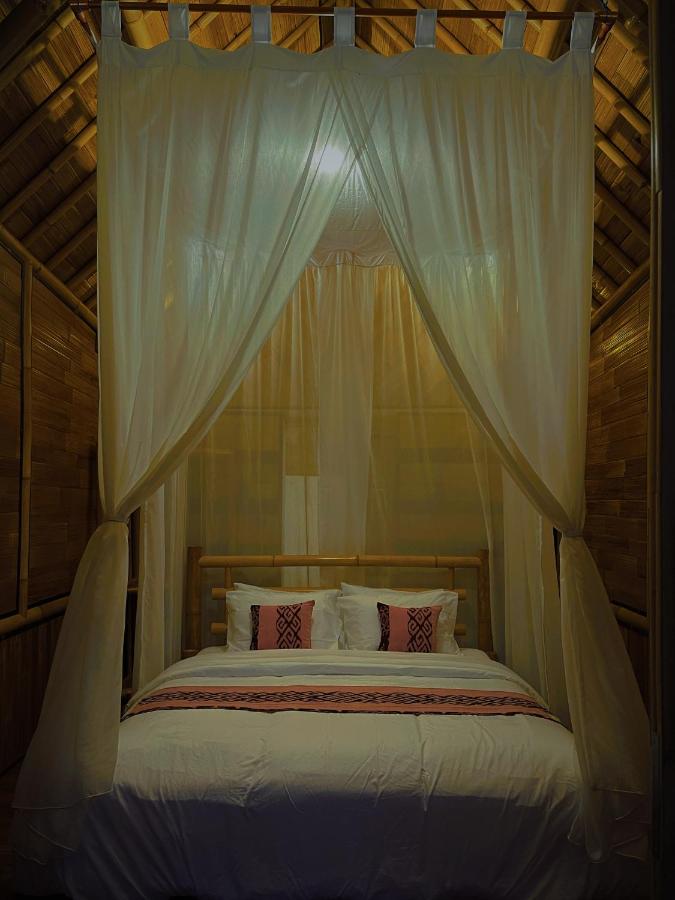 A cozy bed with white linens and patterned pillows is framed by sheer white canopy curtains inside a warm, wooden, thatched-roof room—exuding the inviting charm found in the best family villas in Bali.