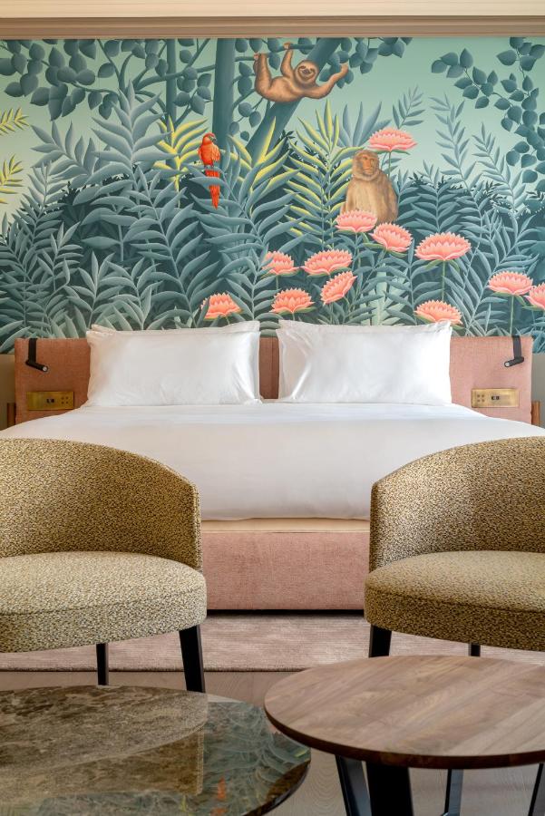 Where to Stay in Madrid: 14 Best Hotels 56 A stylish hotel room with a double bed, two beige armchairs, and a round wooden table—an ideal choice for where to stay in Madrid first time. The headboard wall features a vibrant mural with sloths, a parrot, and lush greenery with pink flowers.