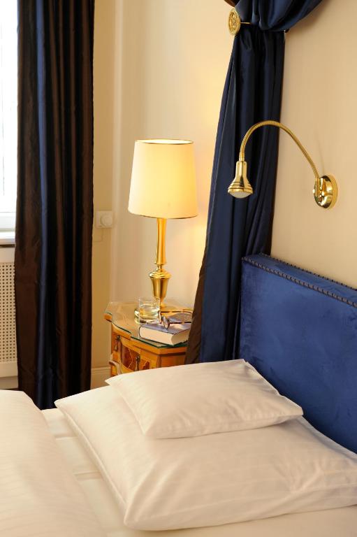 Authentic Boutique Hotels in Munich 17 A neatly made bed with white linens and pillows beside a nightstand holding a glass of water, books, and a lamp—just like the inviting touches found in boutique hotels in Munich. Navy blue drapes and headboard add color to the softly lit room.