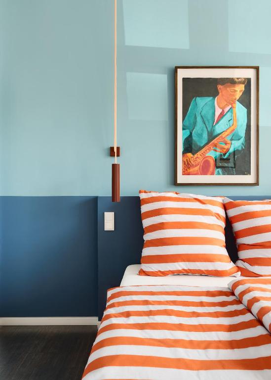 Boutique Hotels in Berlin Kreuzberg 20 A modern bedroom inspired by boutique hotels in Berlin Kreuzberg, with light and dark blue walls, a bed with orange and white striped bedding, a hanging brown pendant light, and a framed painting of a man playing guitar above the bed.