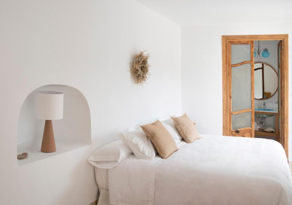 25 Weekend Trips from Madrid Max. 2h Away 70 A minimalist bedroom with a white bed, beige pillows, and a lamp in a wall niche—perfect for restful nights after weekend trips from Madrid. A rustic wooden door leads to a bathroom with a round mirror and blue sink.