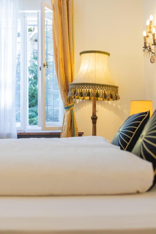 Authentic Boutique Hotels in Munich 9 A cozy bedroom with a neatly made bed, decorative pillows, a vintage floor lamp, gold curtains, and an open window letting in natural light—reminiscent of rooms found in stylish boutique hotels in Munich.