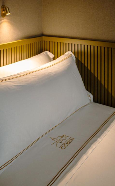 8 Unique Boutique Hotels in Bilbao Spain 2 A neatly made bed with crisp white linens and gold trim, featuring the embroidered logo of Hotel COZIER—one of the top boutique hotels in Bilbao. The bed is set against a light brown paneled wall with a small wall-mounted reading light above.