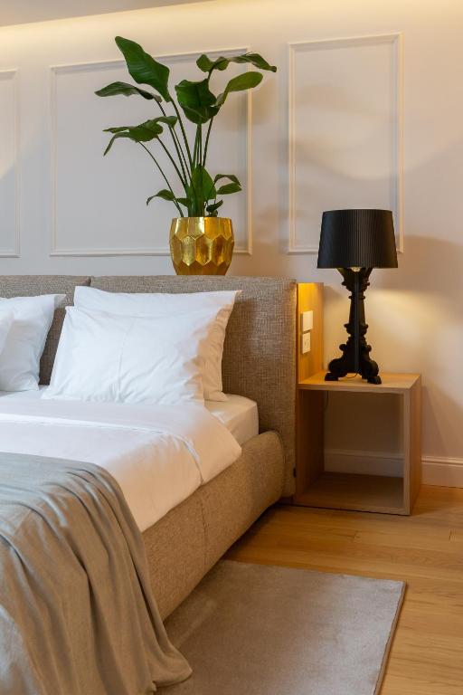 Where to Stay in Split: Hotel & Areas Guide 21 A modern bedroom—perfect inspiration for deciding where to stay in Split—features a beige bed with white bedding, a gray throw blanket, wooden nightstand, black lamp, and gold plant pot with green leaves against a paneled wall.