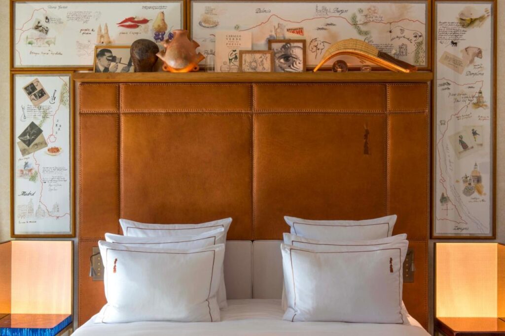Where to Stay in Madrid: 14 Best Hotels 24 A neatly made bed with four white pillows sits before a tan leather headboard. The wall behind features sketches, maps, and small objects—an inviting look for anyone seeking where to stay in Madrid first time.