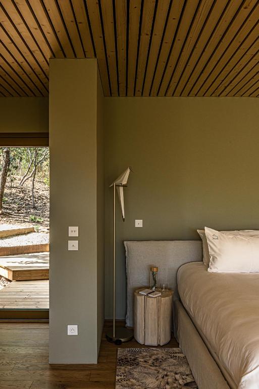 11 Unique Places to Stay in Provence 24 A modern bedroom with a green accent wall, a large bed with white bedding, a wooden bedside table, and a floor lamp overlooks a wooden deck and trees—one of the unique places to stay in Provence.