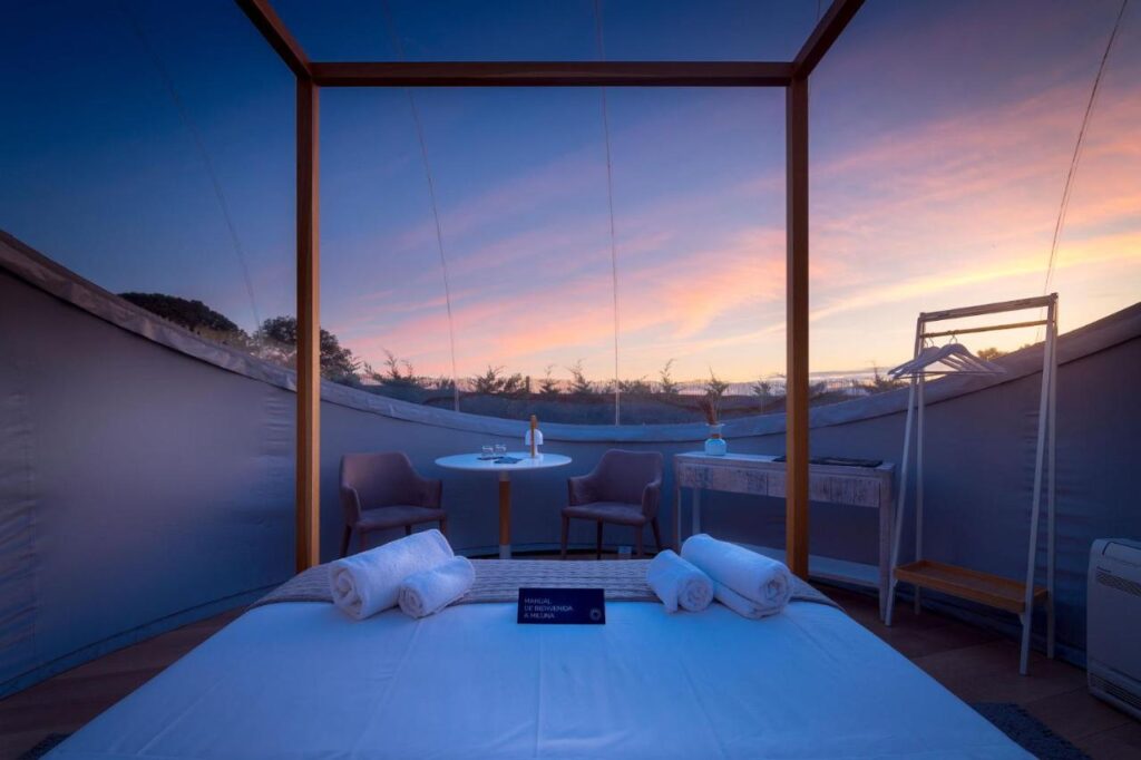 25 Weekend Trips from Madrid Max. 2h Away 20 A cozy, transparent tent room with a bed, chairs, and table—perfect for weekend trips from Madrid—offers a scenic sunset view through clear walls. Rolled towels rest on the bed, and soft lighting creates a relaxing atmosphere.