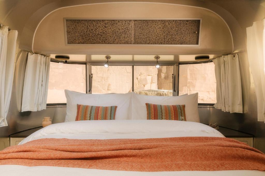 9 Best Hotels in Saudi Arabia: Unique Stays Across the Country 24 A cozy bed with white linens, colorful striped pillows, and an orange throw blanket inside a camper van, reminiscent of the comfort found in the Best Hotels in Saudi Arabia, with sunlight streaming through large windows and white curtains.