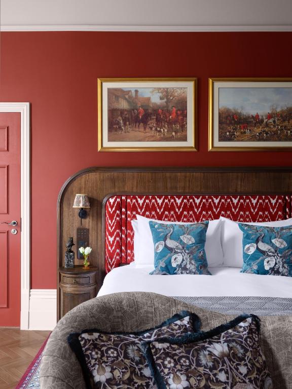15 Best Boutique Hotels in London, UK 30 A stylish bedroom inspired by boutique hotels in London, featuring a red accent wall, patterned headboard, two framed paintings above the bed, blue floral pillows, and a round bedside table with a lamp and vase of flowers.