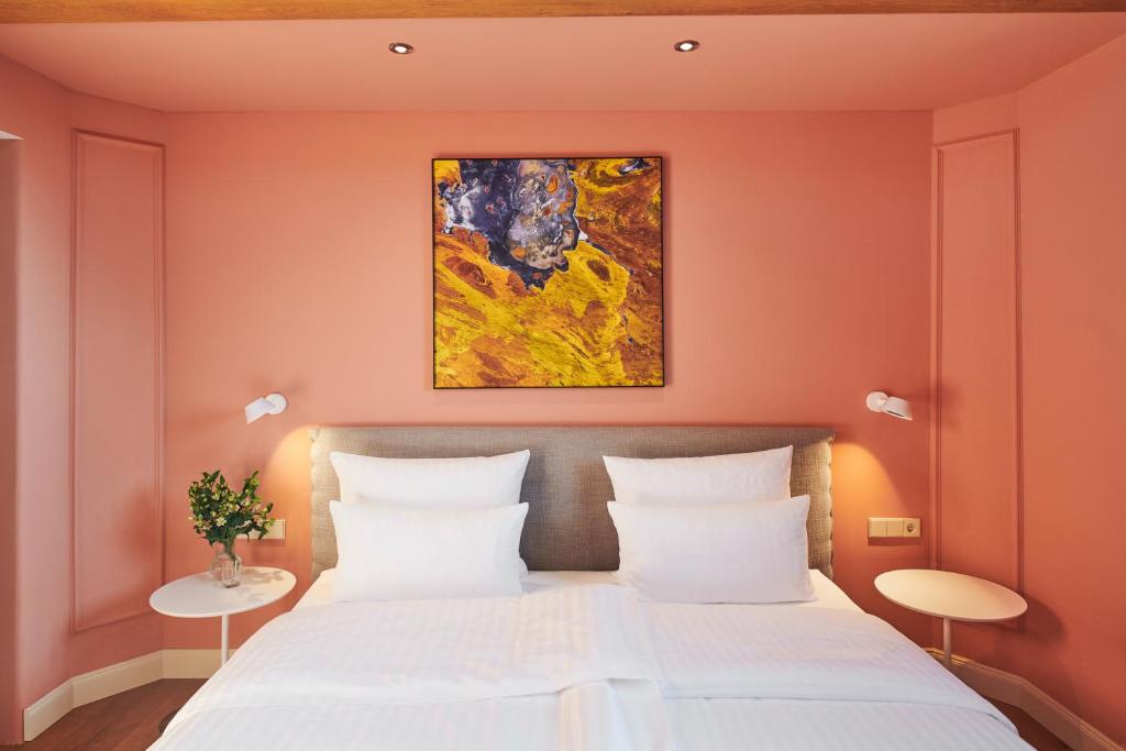 Authentic Boutique Hotels in Munich 24 A neatly made bed with white linens sits against a peach-colored wall, evoking the style of boutique hotels in Munich. Above the bed hangs an abstract painting, while round side tables with lamps and a plant complete the cozy, modern room.