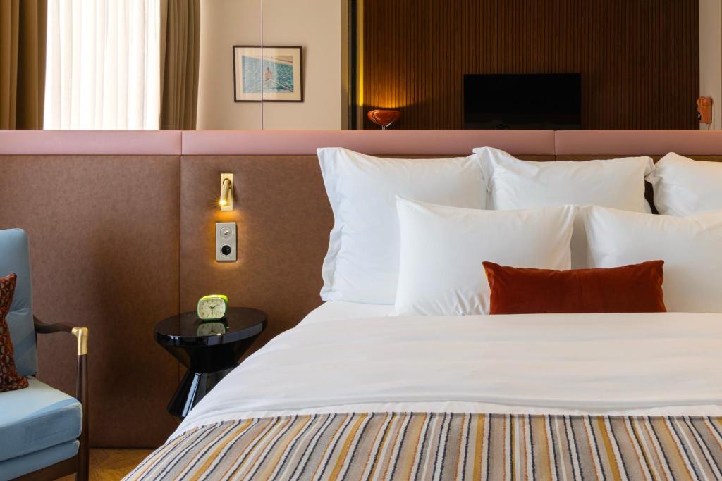 11 Unique Hotels in Georgia You Can't Miss! 18 A neatly made bed with white pillows and a rust-colored cushion stands beside a small black side table with a retro clock and lamp. A blue armchair and wall art add charm to the modern, well-lit roomāperfect for unique hotels in Georgia.