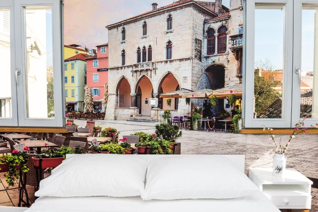 8 TOP Boutique Hotels in Split 13 A bed with white pillows and bedding stands in front of large windows, offering a vibrant view of a European plaza—perfect inspiration for those searching Where to Stay in Split amid outdoor cafes, colorful buildings, and historic architecture.