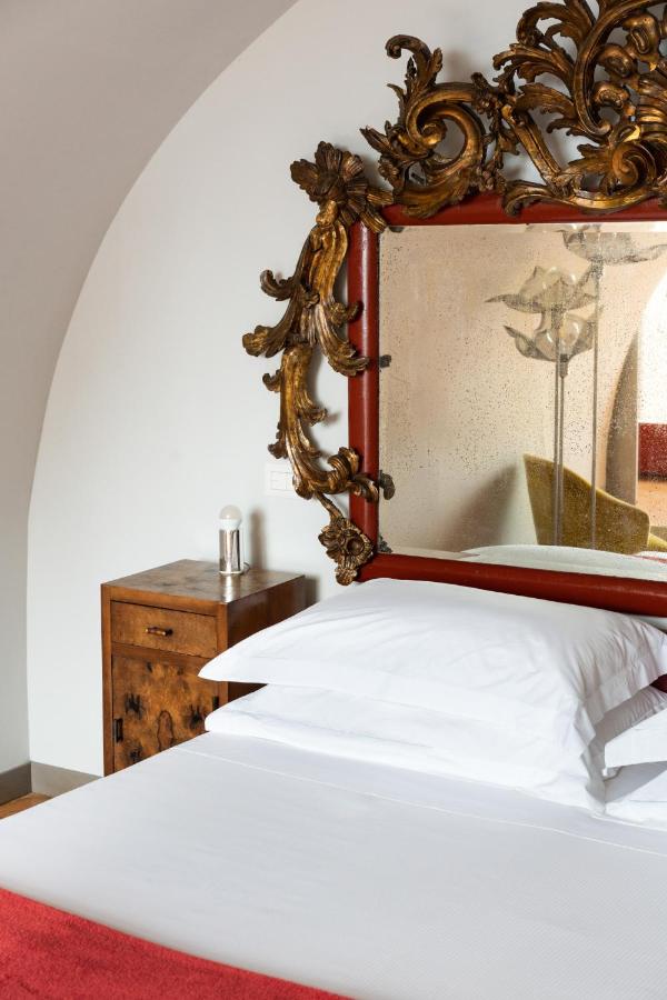 Boutique Hotels on the Amalfi Coast 6 A neatly made bed with white pillows and sheets sits beside a wooden nightstand with a lamp—reminiscent of the elegant decor found in boutique hotels on the Amalfi Coast. Above the bed, a large ornate mirror boasts an intricate gold frame.