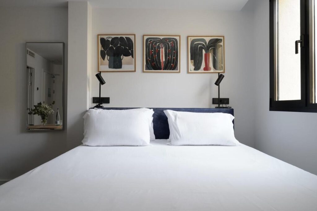 Where to Stay in Madrid: 14 Best Hotels 28 A neatly made bed with white sheets and two pillows is centered in a modern bedroom—an ideal inspiration for those searching where to stay in Madrid first time. Above the bed, three abstract paintings add character, with natural light streaming through the window.