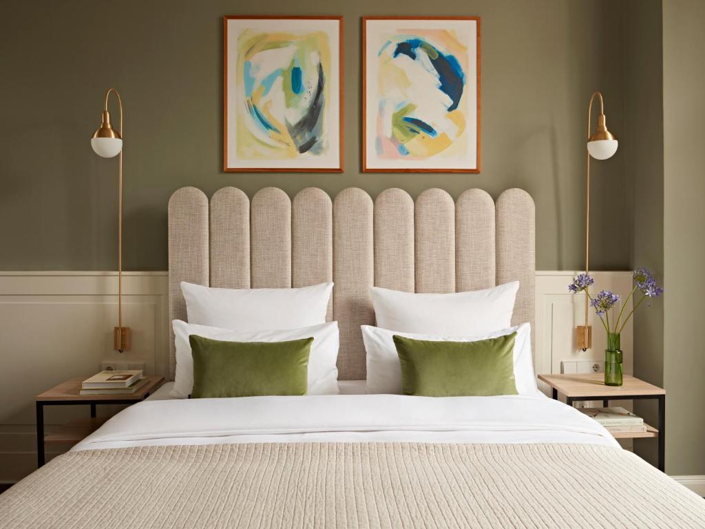 Boutique Hotels in Berlin Kreuzberg 16 A neatly made bed with a beige scalloped headboard, white bedding, and green accent pillows evokes the style of boutique hotels in Berlin Kreuzberg. Two abstract paintings hang above, with wall-mounted lamps and vases of flowers on bedside tables.