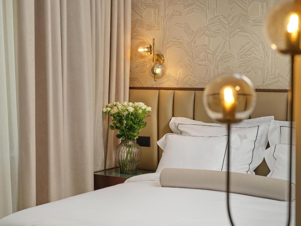 8 TOP Boutique Hotels in Split 30 A cozy bedroom reminiscent of boutique hotels in Split, featuring a neatly made bed, white linens, a beige bolster, a glass vase of white roses on the nightstand, modern wall sconces, and soft beige curtains.