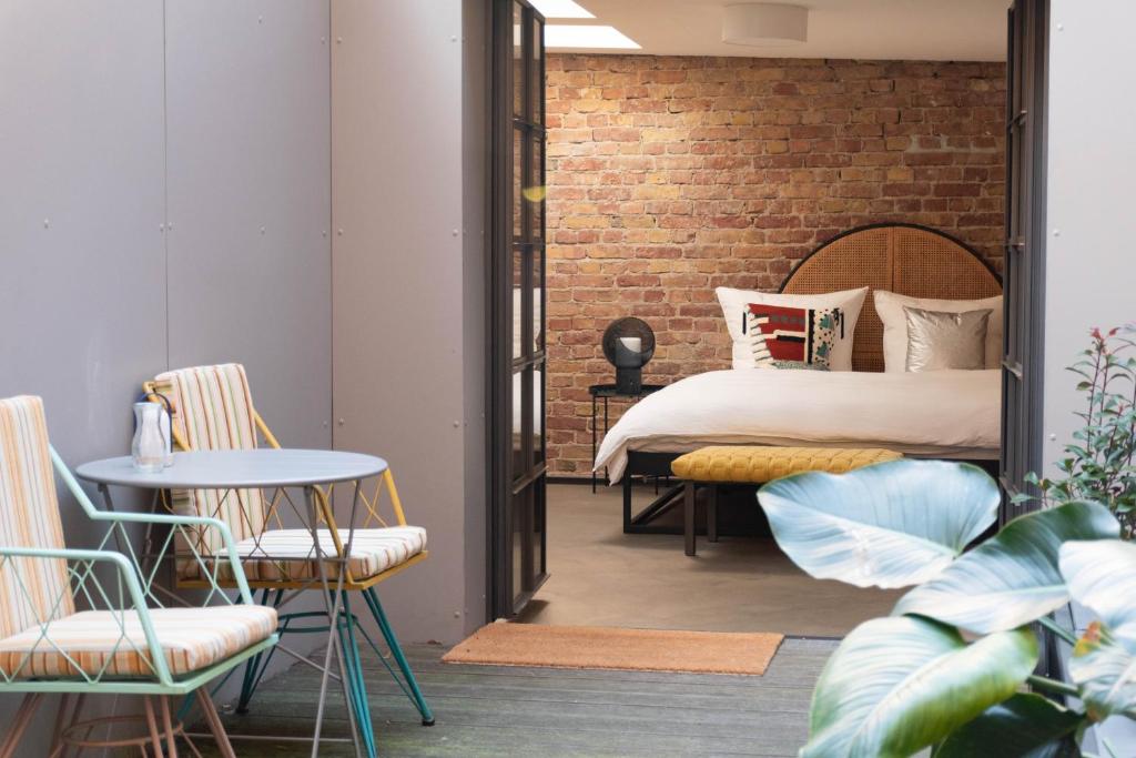 Boutique Hotels in Berlin Kreuzberg 26 A cozy bedroom with exposed brick walls and a large bed is seen through open double doorsāa vibe often found in boutique hotels in Berlin Kreuzberg. Outside, a small patio features a round table, striped chairs, and a potted plant in the foreground.