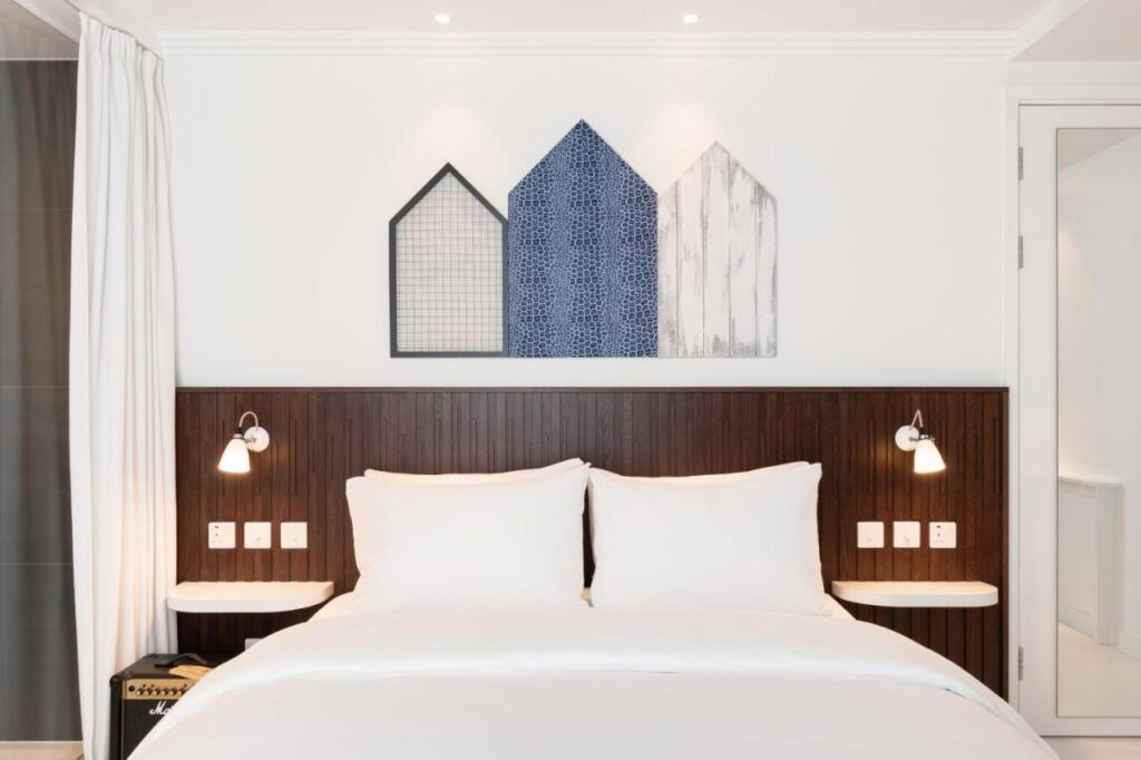 15 Best Boutique Hotels in London, UK 19 A modern, minimalistic bedroom inspired by the best boutique hotels London offers, featuring a double bed with white bedding, dark wooden headboard, wall-mounted reading lights, and geometric house-shaped art above the bed.