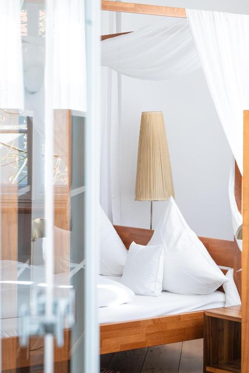 FAIRYTALE Castle Hotels in Bavaria 9 A bright, modern bedroom with white bedding, fluffy pillows, a wooden canopy bed frame, and white curtains evokes the elegance found in castle hotels in Bavaria. A wooden nightstand and tall pleated beige lampshade sit next to the bed.
