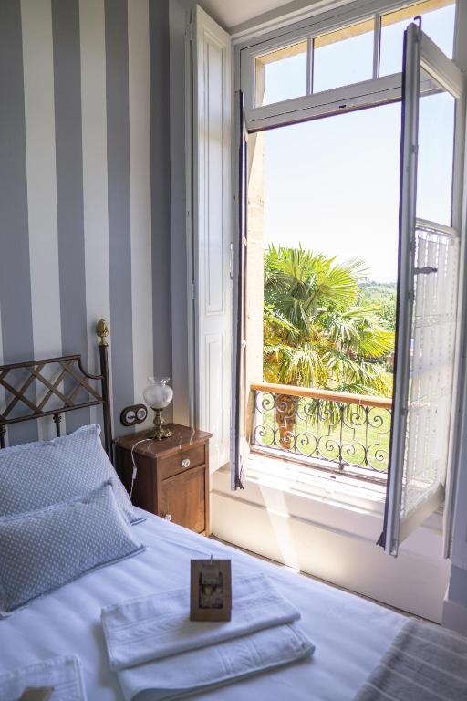 A sunny bedroom reminiscent of boutique hotels in Asturias, featuring a striped wall, a neatly made bed with white linens, a wooden nightstand, and an open window overlooking palm trees and lush greenery.
