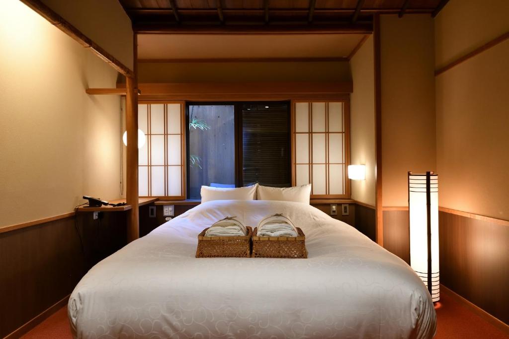 7 UNIQUE Ryokans in Gion Kyoto 23 A cozy Japanese-style bedroom inspired by ryokans in Gion, Kyoto, with a large bed, white bedding, two baskets with folded robes, shoji screen windows, wooden walls, and a tall floor lamp emitting soft light.