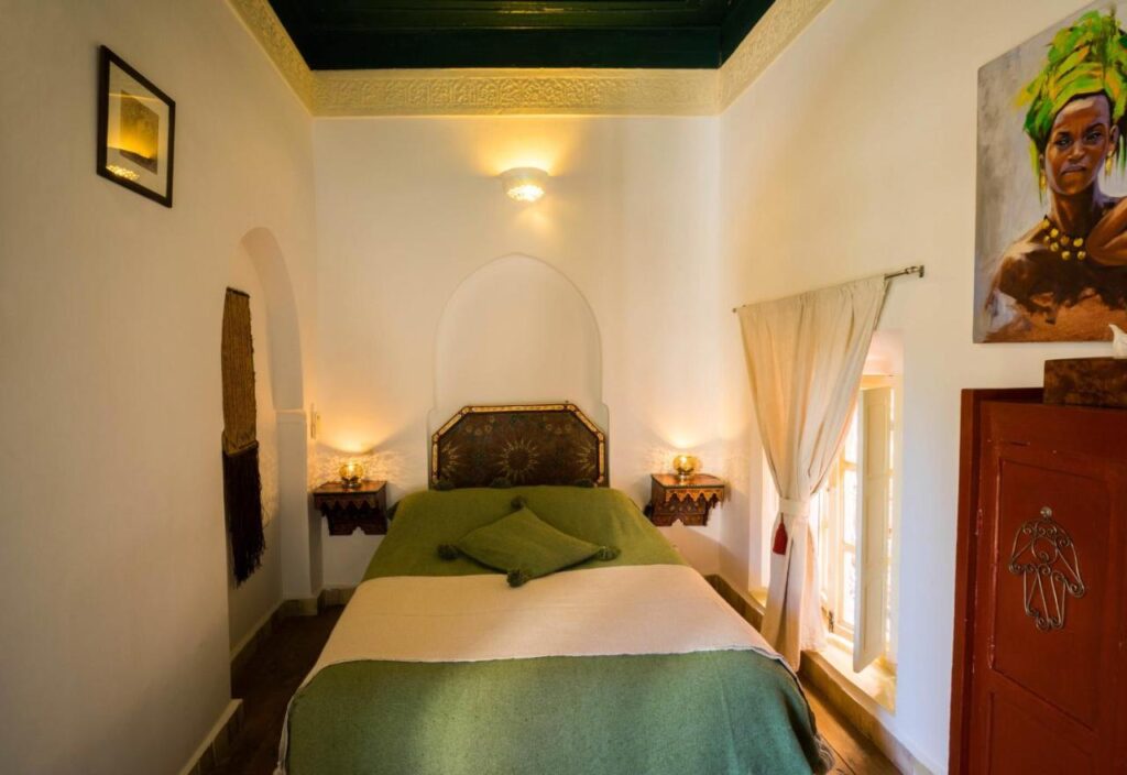 12 Charming Budget Riads in Marrakech 13 A cozy bedroom, inspired by budget riads in Marrakech, features a green and beige bedspread, triangle-arranged pillows, wall-mounted lamps, white walls, a dark green ceiling, artwork, and sheer curtains letting in natural light.