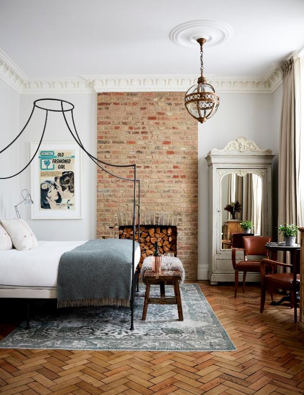 15 Best Boutique Hotels in London, UK 34 A stylish bedroom inspired by boutique hotels in London, featuring a canopy bed, exposed brick fireplace with stacked wood, vintage wardrobe, cozy seating area, parquet floor, neutral tones, framed art, and abundant natural light for an elegant ambiance.