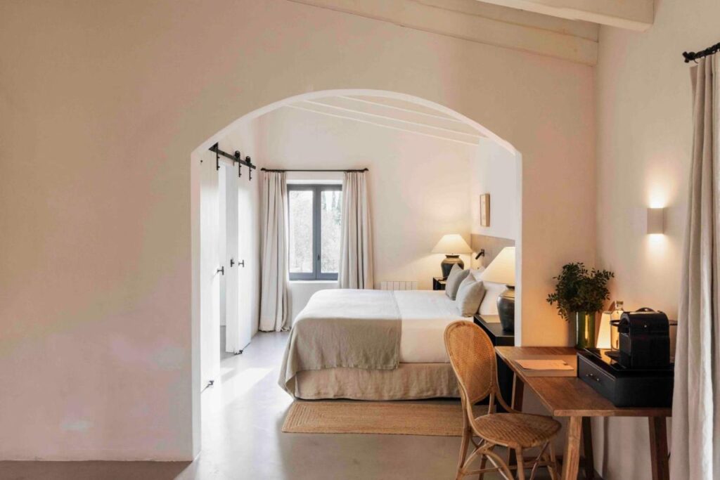Unique Agroturismos in Mallorca 33 A cozy bedroom with neutral tones features a double bed, a wooden desk with a chair and lamp, and a window with white curtains letting in daylight—perfect for relaxing after exploring agroturismos in Mallorca. Plants and soft lighting create a warm, inviting atmosphere.