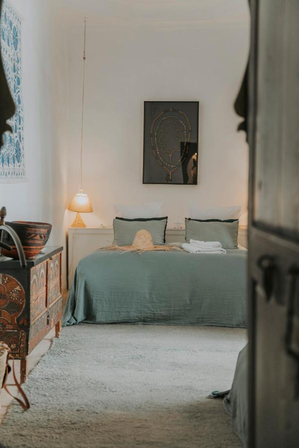 12 Charming Budget Riads in Marrakech 49 A cozy bedroom in the style of budget riads in Marrakech, with a neatly made bed, greenish-gray bedding, and two pillows. Soft bedside lighting creates a warm atmosphere, complemented by a decorative chest and framed artwork.