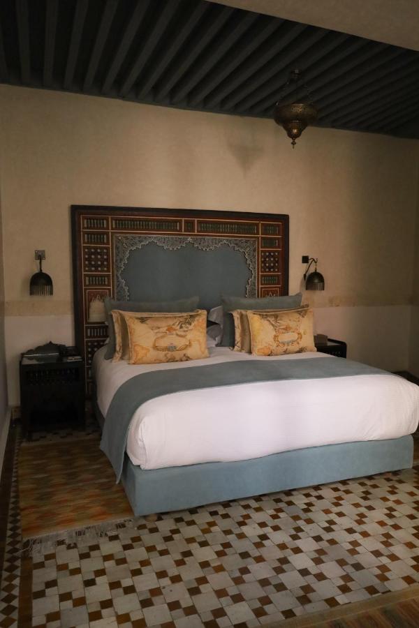 7 UNIQUE Riads in Fez You Won't Forget 18 A neatly made bed with a decorative headboard, blue and beige bedding, and patterned pillows stands in the center of a room with mosaic tile flooring and ornate hanging lamps, reflecting the elegance of riads in Fez.